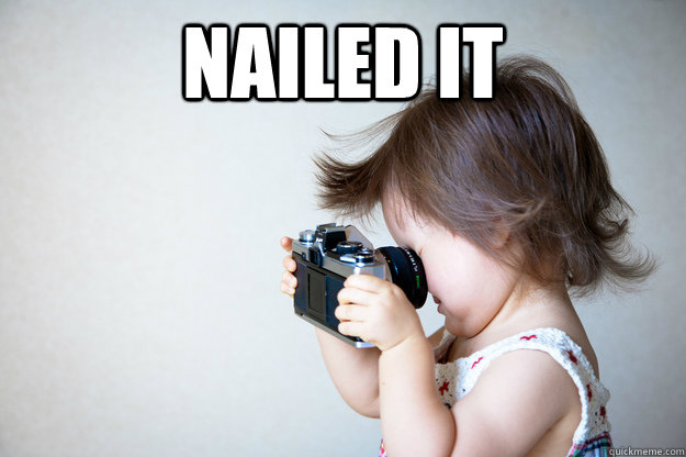 A very young child holds a camera backwards, staring confidently into the lens instead of the viewfinder. The caption says Nailed It.